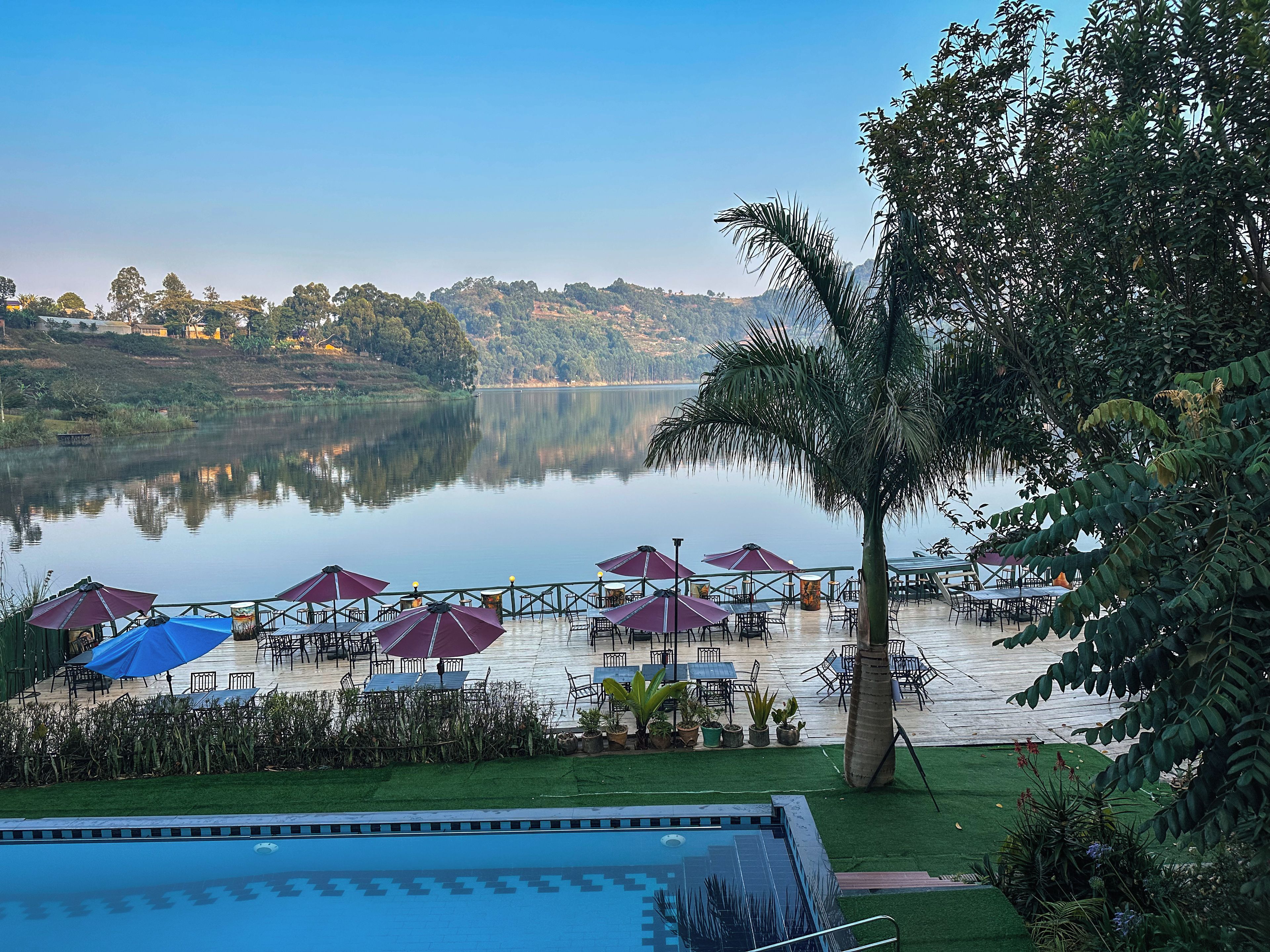 Un espacio al aire libre junto al lago con sombrillas y mesas, rodeado de vegetación y una piscina.