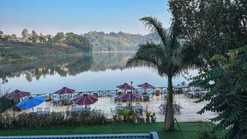 Un espacio al aire libre junto al lago con sombrillas y mesas, rodeado de vegetación y una piscina.