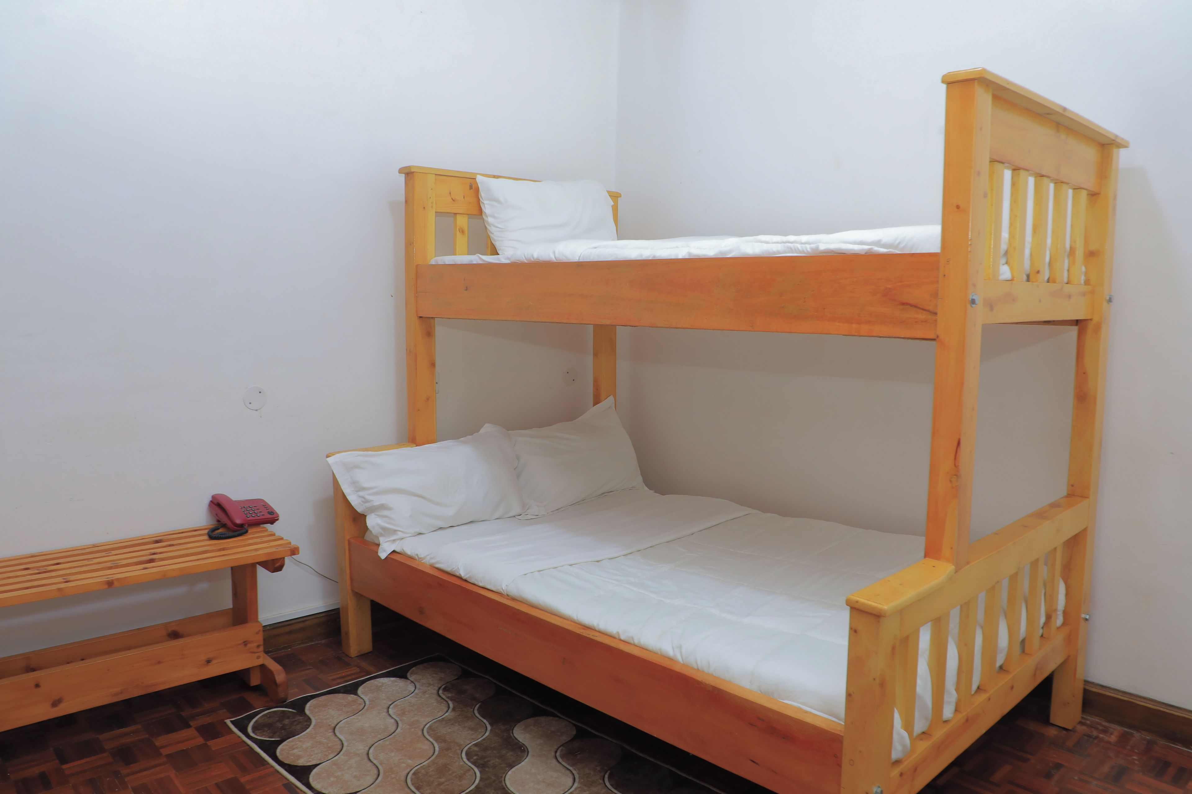 Una cama litera con una cama inferior y una individual, un pequeño banco de madera y un tapete estampado sobre un suelo de madera.