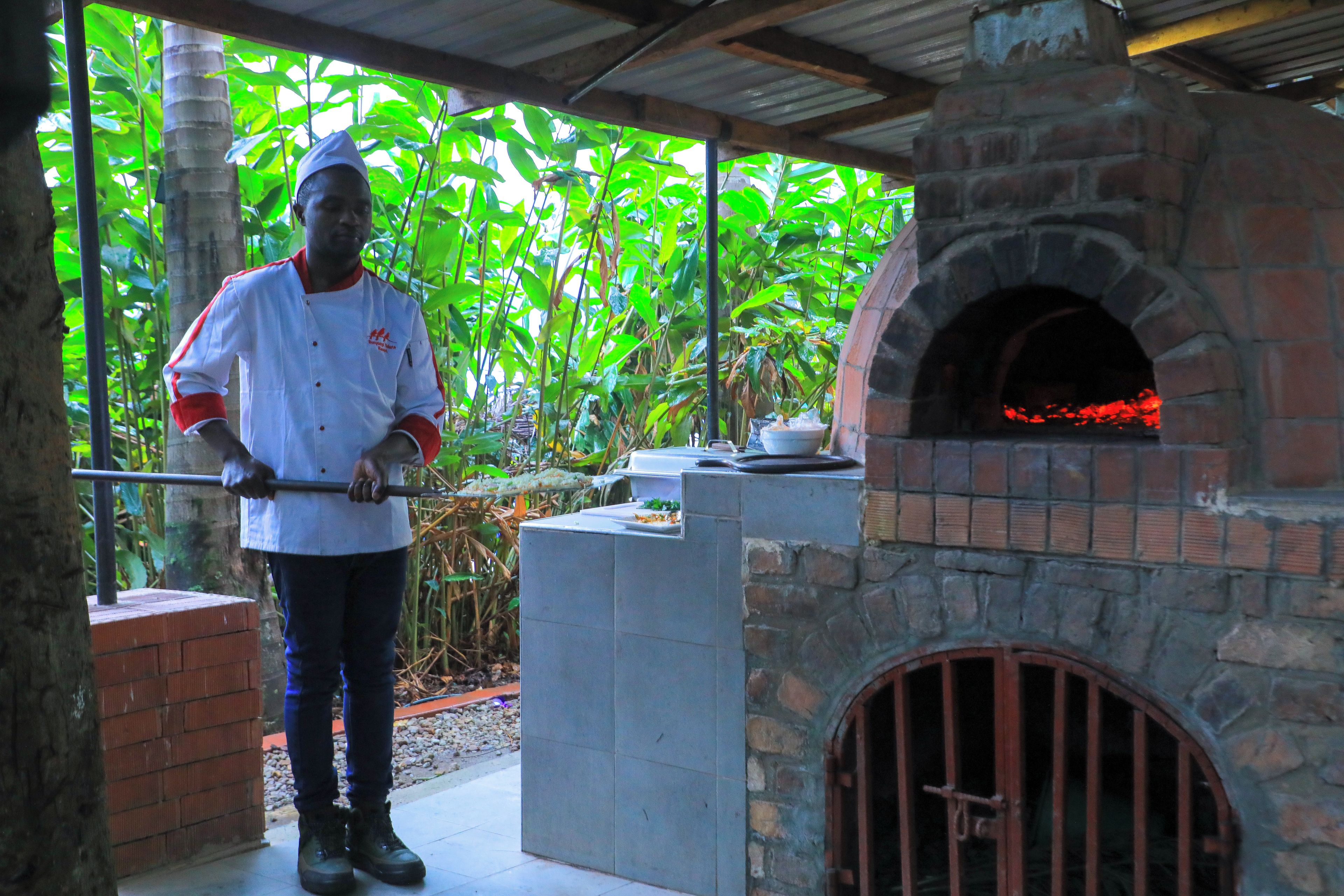 A chef standing in front of a stone oven with a grill in a covered outdoor area.