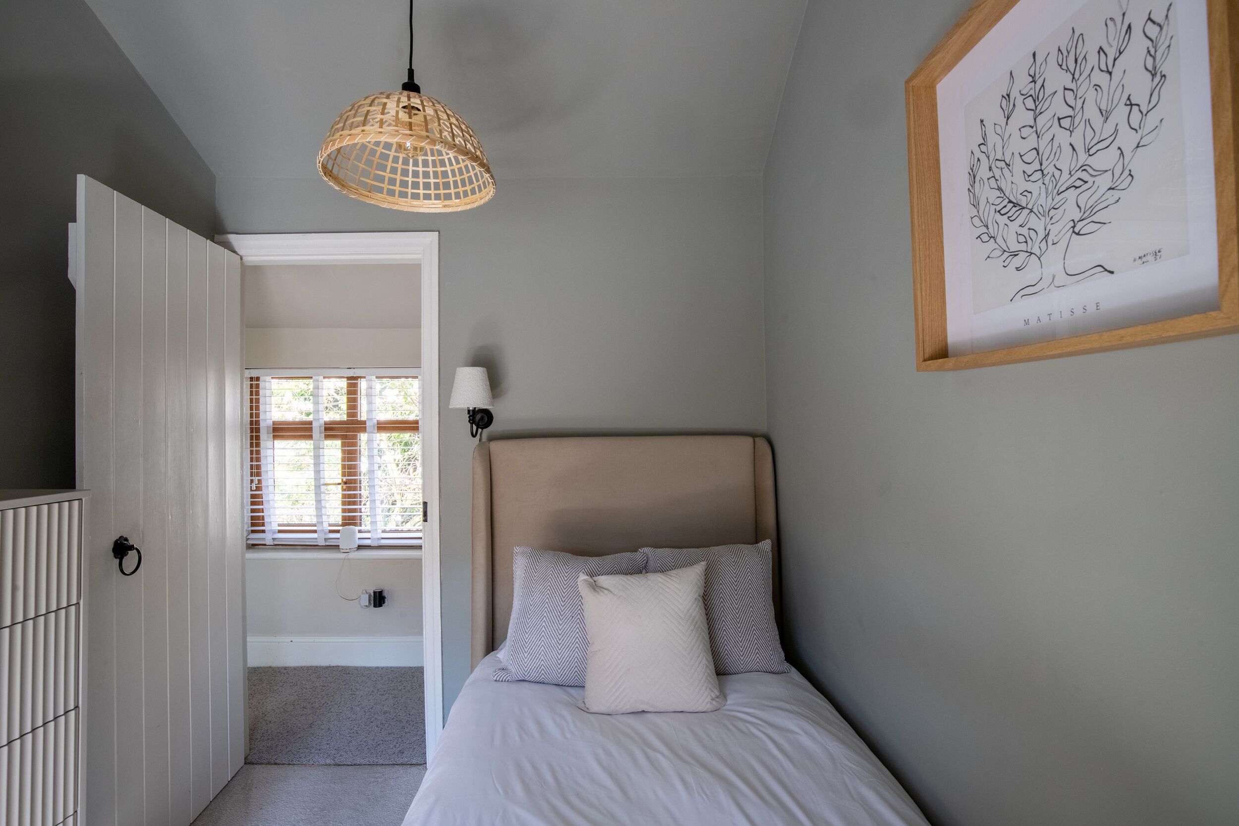 Ein Schlafzimmer mit einem Bett, einer Pendelleuchte, einem gerahmten Kunstwerk und einem Fenster mit Jalousien.