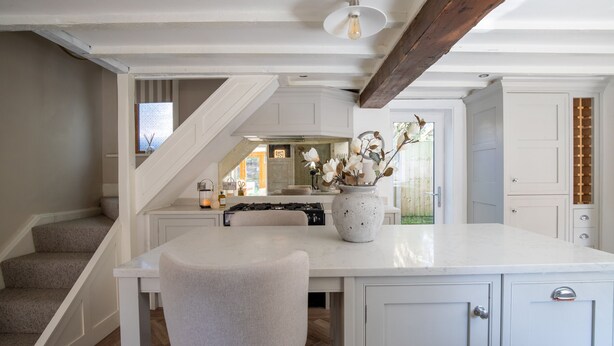 A kitchen with a central island, white cabinetry, a wooden staircase, and a view of the outside. - Rainbow Cottage (Ormskirk)
