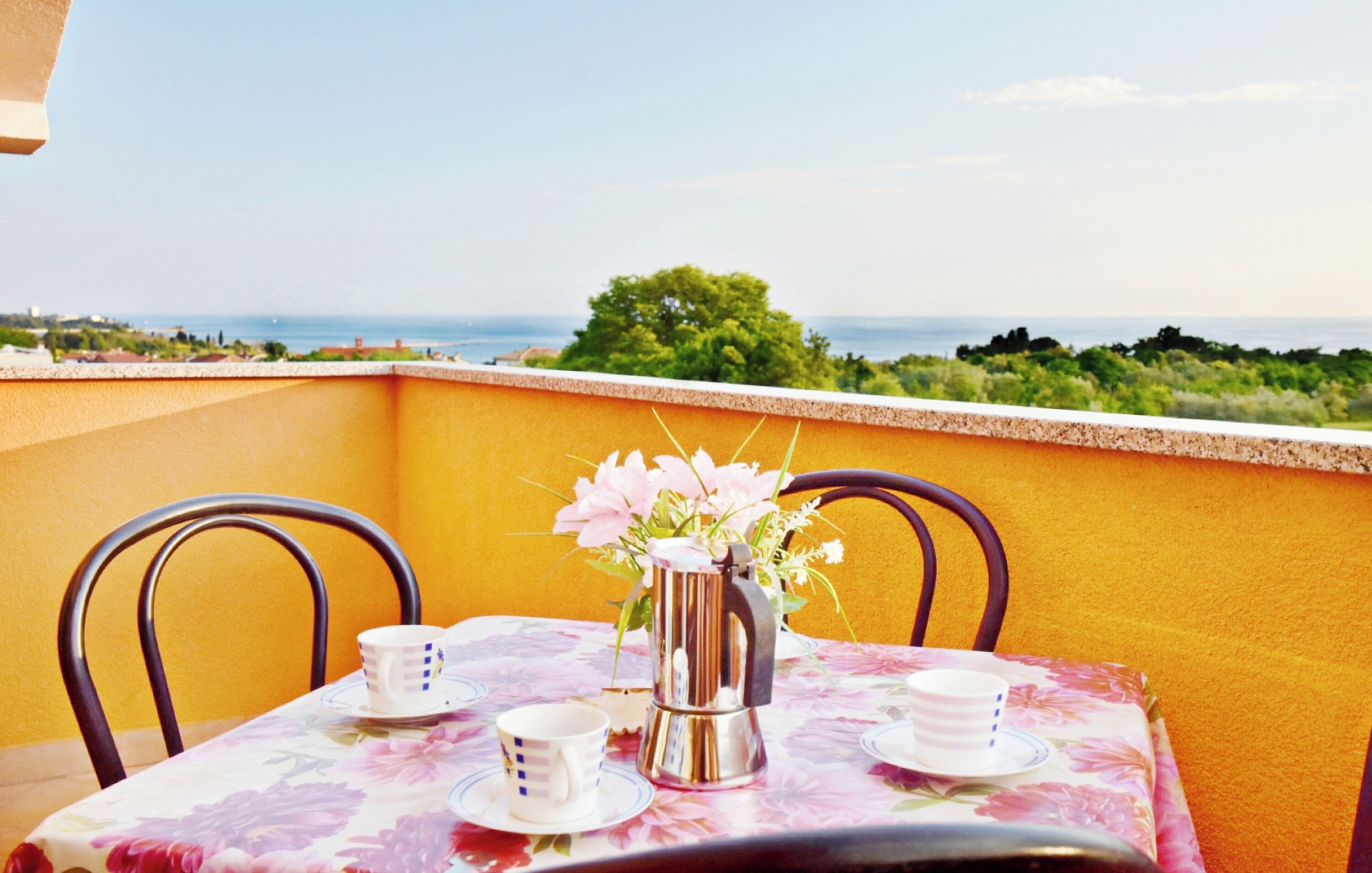 A table set with cups and a flower vase on a balcony with a view of the sea.