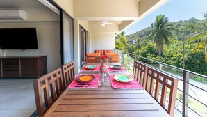 A balcony with a wooden table set for four, equipped with colorful placemats, plates, and cutlery. A flat-screen TV is mounted on the wall to the left. - Short Walk to Beach & Town · Top Level · Solana (Huatulco)