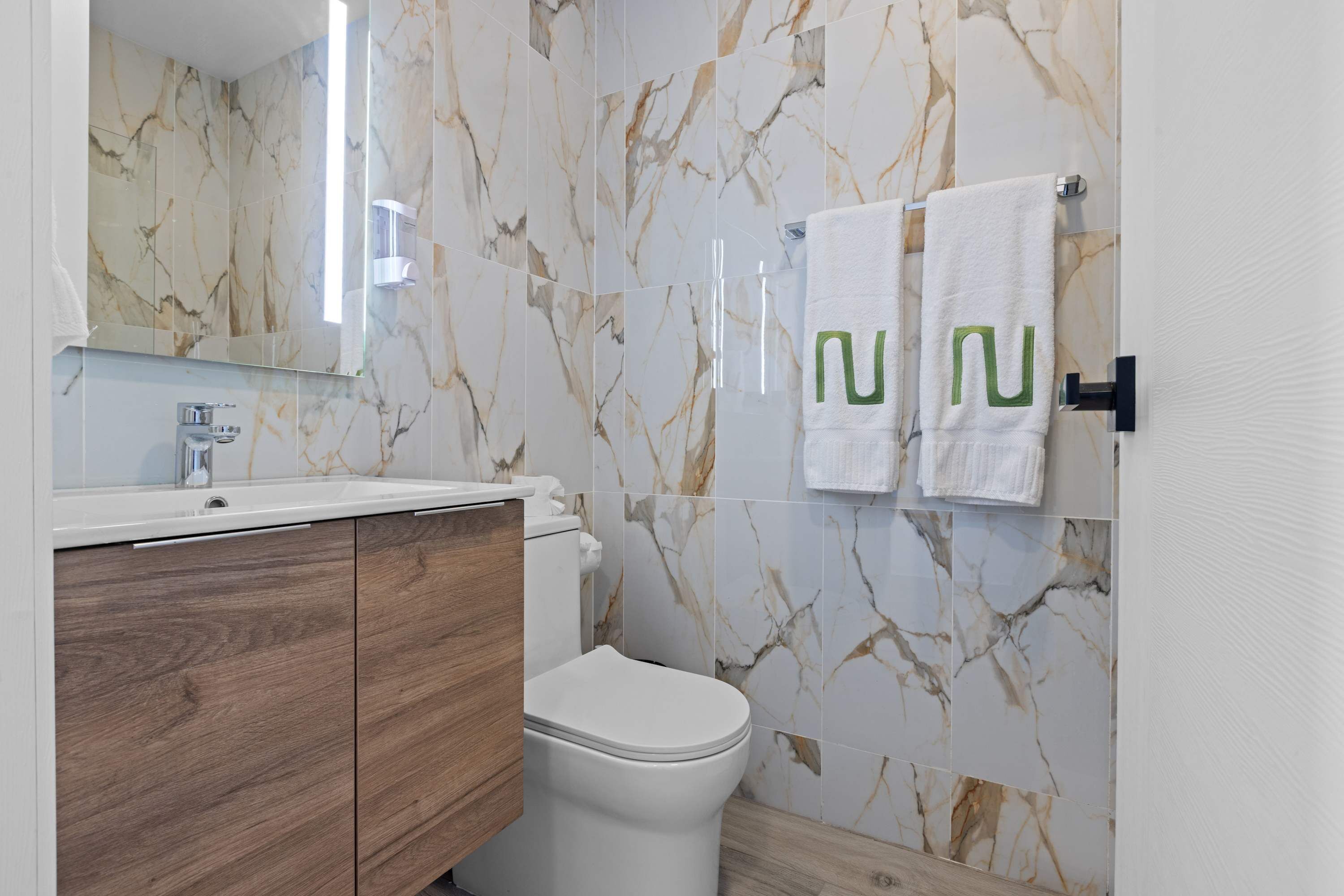 Ein modernes Badezimmer mit Marmorwänden, einem Holzkosmetikschrank, einer weißen Toilette und zwei Handtüchern mit grünem Aufdruck.