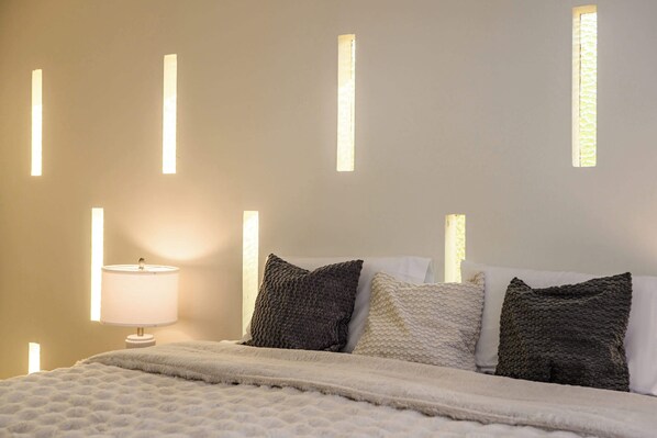 A neatly made bed with a white duvet and several pillows, a bedside lamp, and a wall with vertical light panels. - Modern 5BR Villa w AC Fast Wifi Near Crypto (Monterey Park)