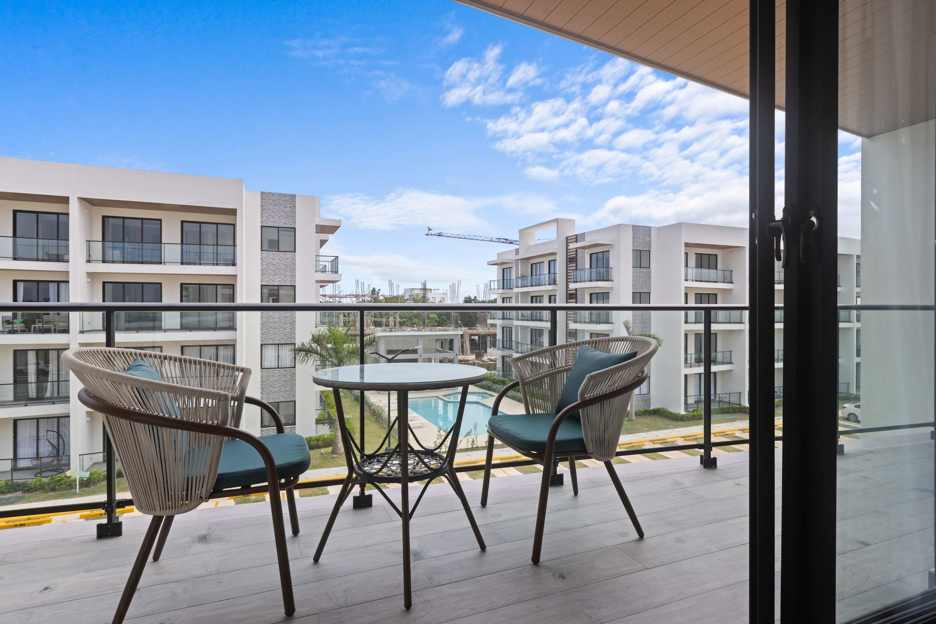 A balcony with wicker chairs and a small table, overlooking a swimming pool and modern buildings.