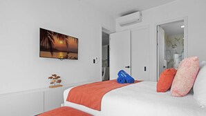 A bedroom with a bed, a painting of a sunset, a white air conditioning unit, and a door leading to another room. - NOK Elegant 2BR Apt by Vistacana (Punta Cana)