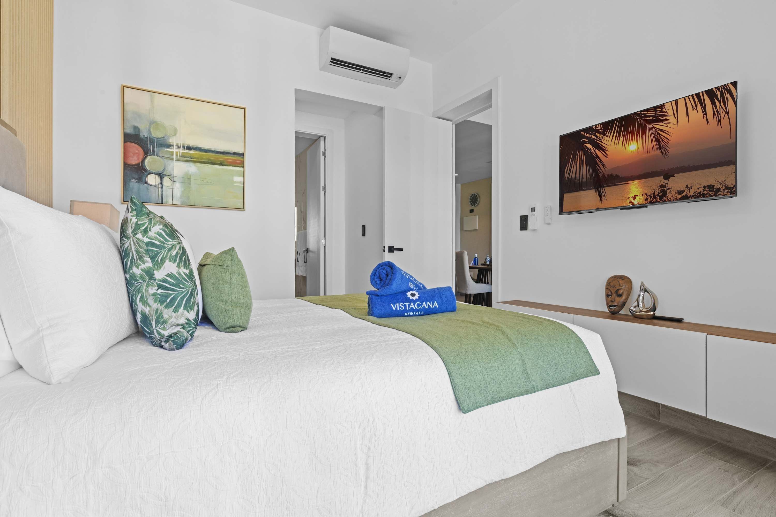 A hotel room with a bed, a painting of a sunset, a wall-mounted air conditioner, and a door leading to another room.