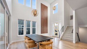 A spacious dining area with a dark wooden table and light brown chairs, large windows, and a staircase leading to another level. - Ski Chalet Chic Retreat With Hot Tub (Park City)