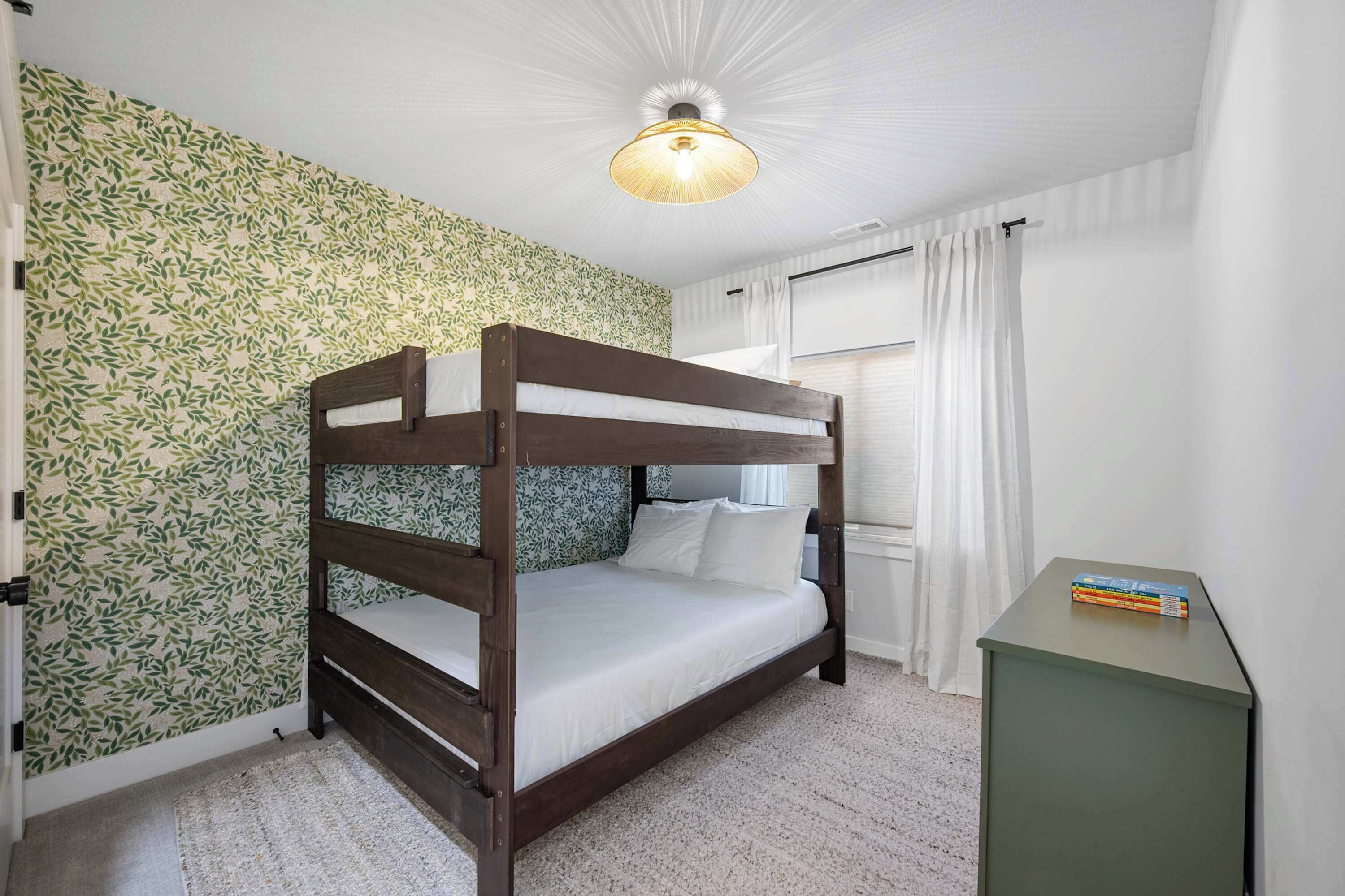 Uma cama beliche com estrutura de madeira e roupa de cama branca, uma pequena mesa de cabeceira com livros, e uma parede com padrão de folhas.