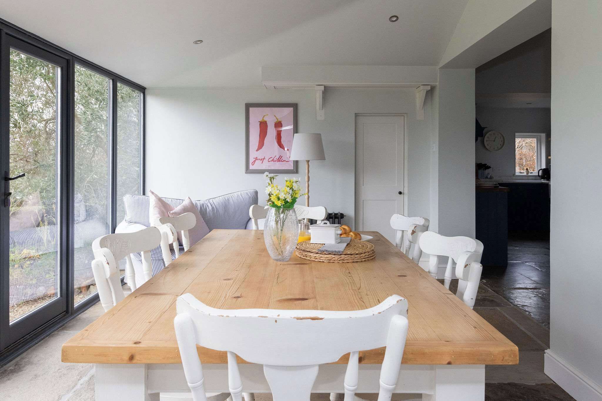 Un coin repas avec une table en bois, des chaises blanches, un vase de fleurs et un tableau encadré au mur.
