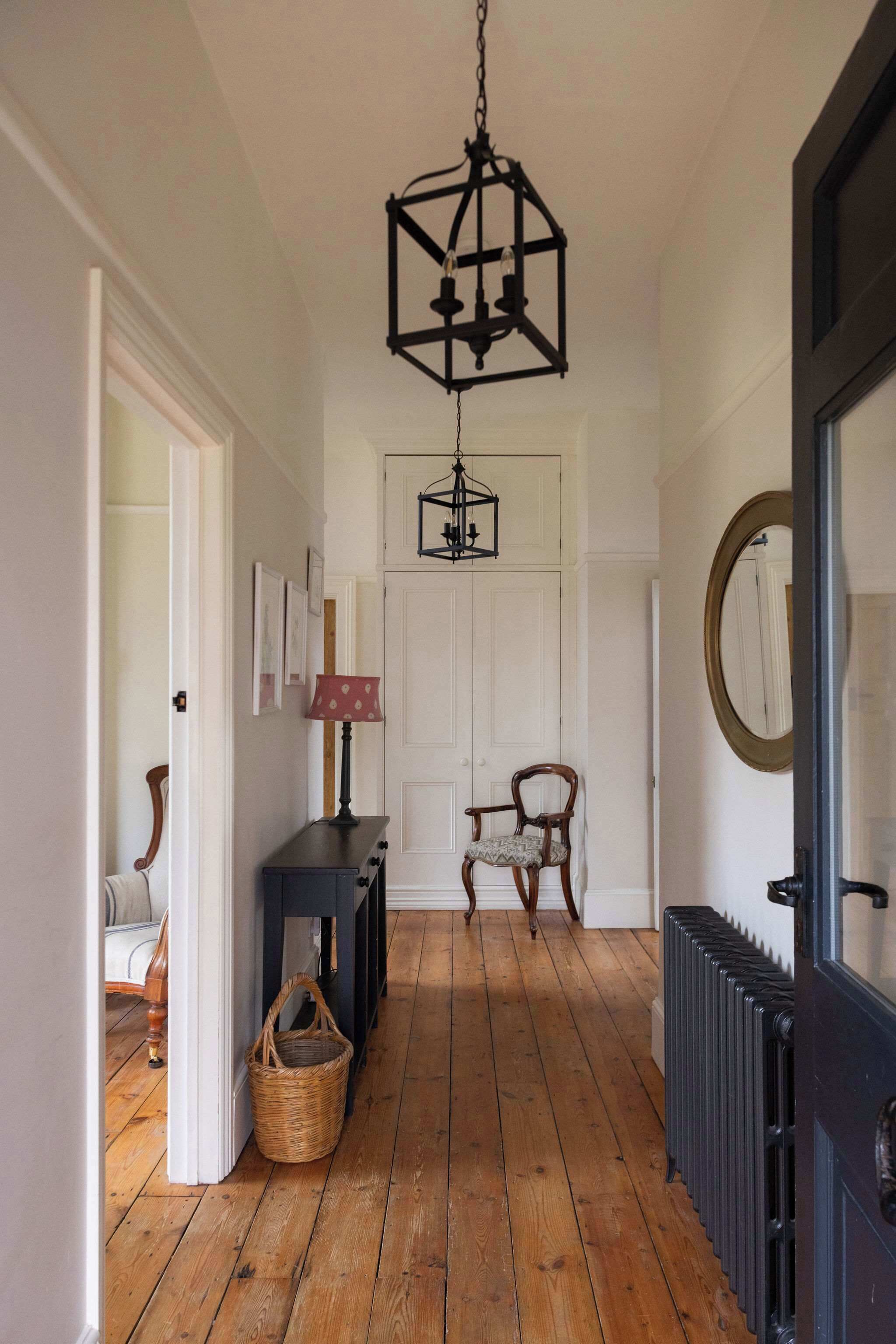 A hallway with wooden flooring, a black hanging light fixture, a white door, and a chair.