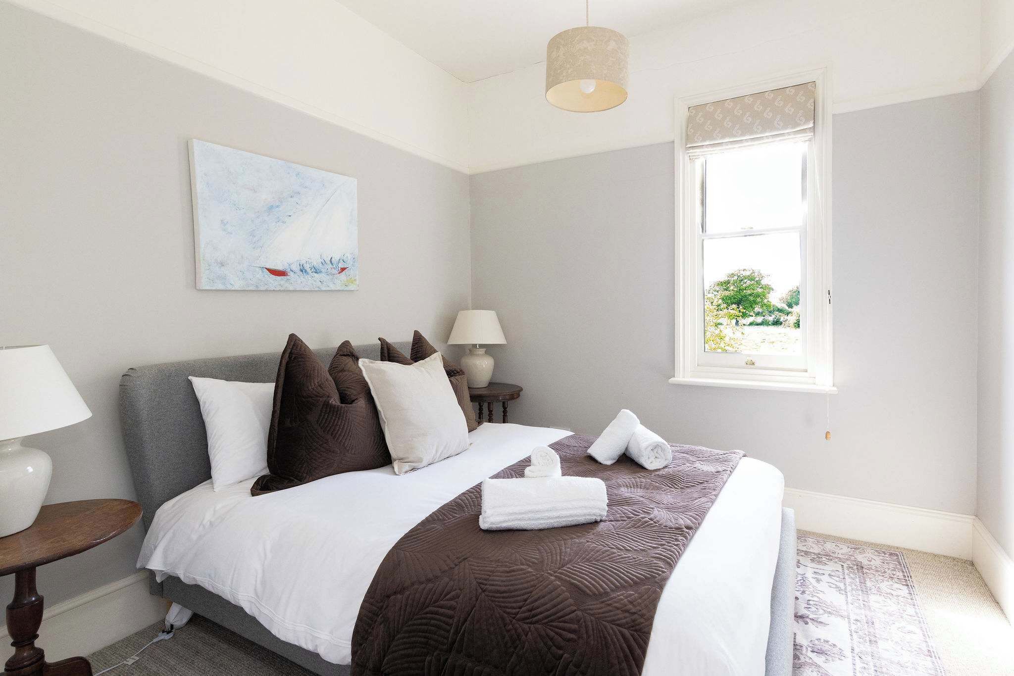 Ein ordentlich bezogenes Bett mit weißer und brauner Bettwäsche, ein Nachttisch mit Lampe, ein Bild an der Wand und ein Fenster mit Blick auf Grünflächen.