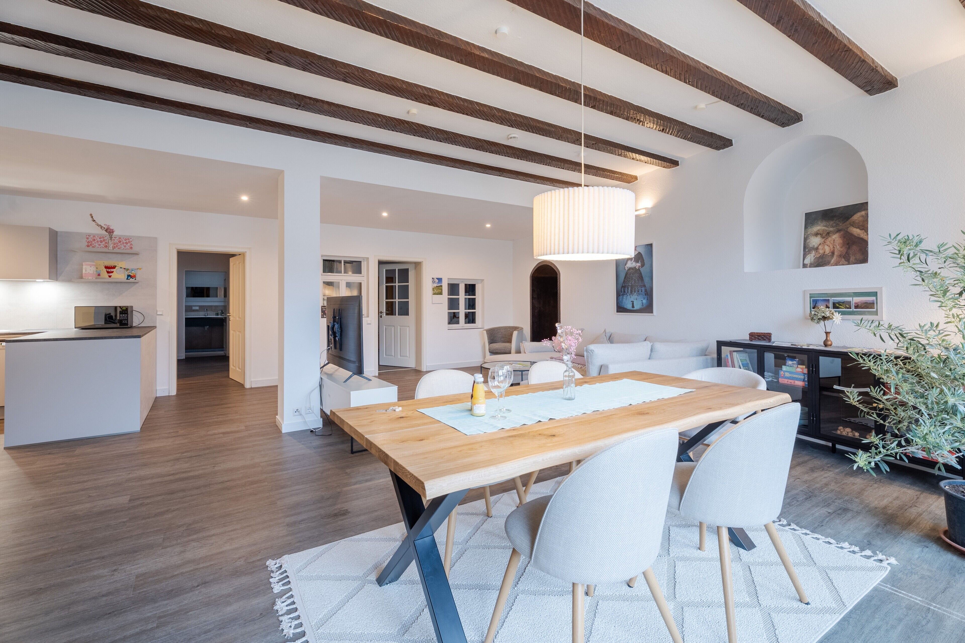 Un vaste espace de vie comprenant une table à manger en bois, des chaises blanches, un canapé et, au fond, une cuisine.