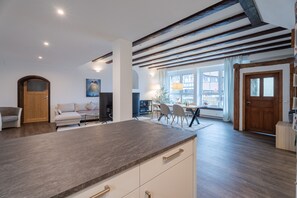 Moderni keittiö, jossa on suuri keittiösaareke, puupalkit katossa sekä ruokailutila, jossa on tuoleja ja pöytä.