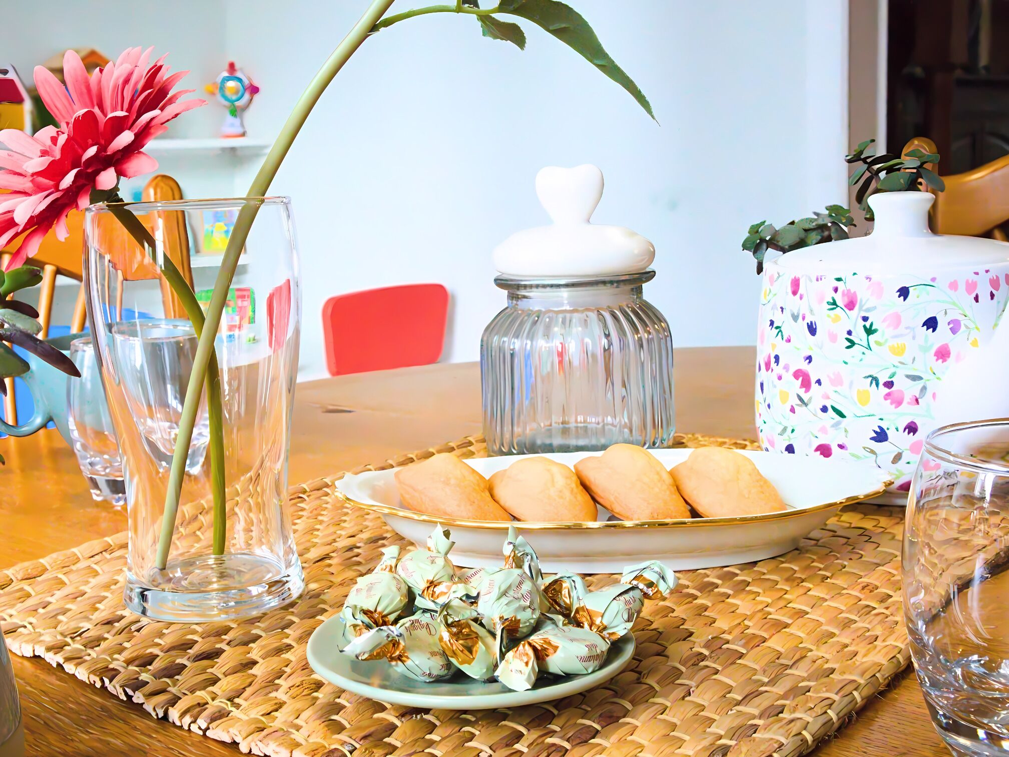 A table with a woven placemat, a glass jar, a floral-patterned container, a plate with cookies, and a plate with wrapped candies.