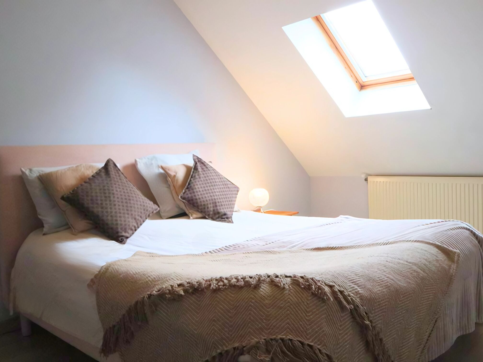 Une chambre avec un plafond en pente, un puits de lumière, un lit avec des oreillers, une lampe de chevet et un radiateur.