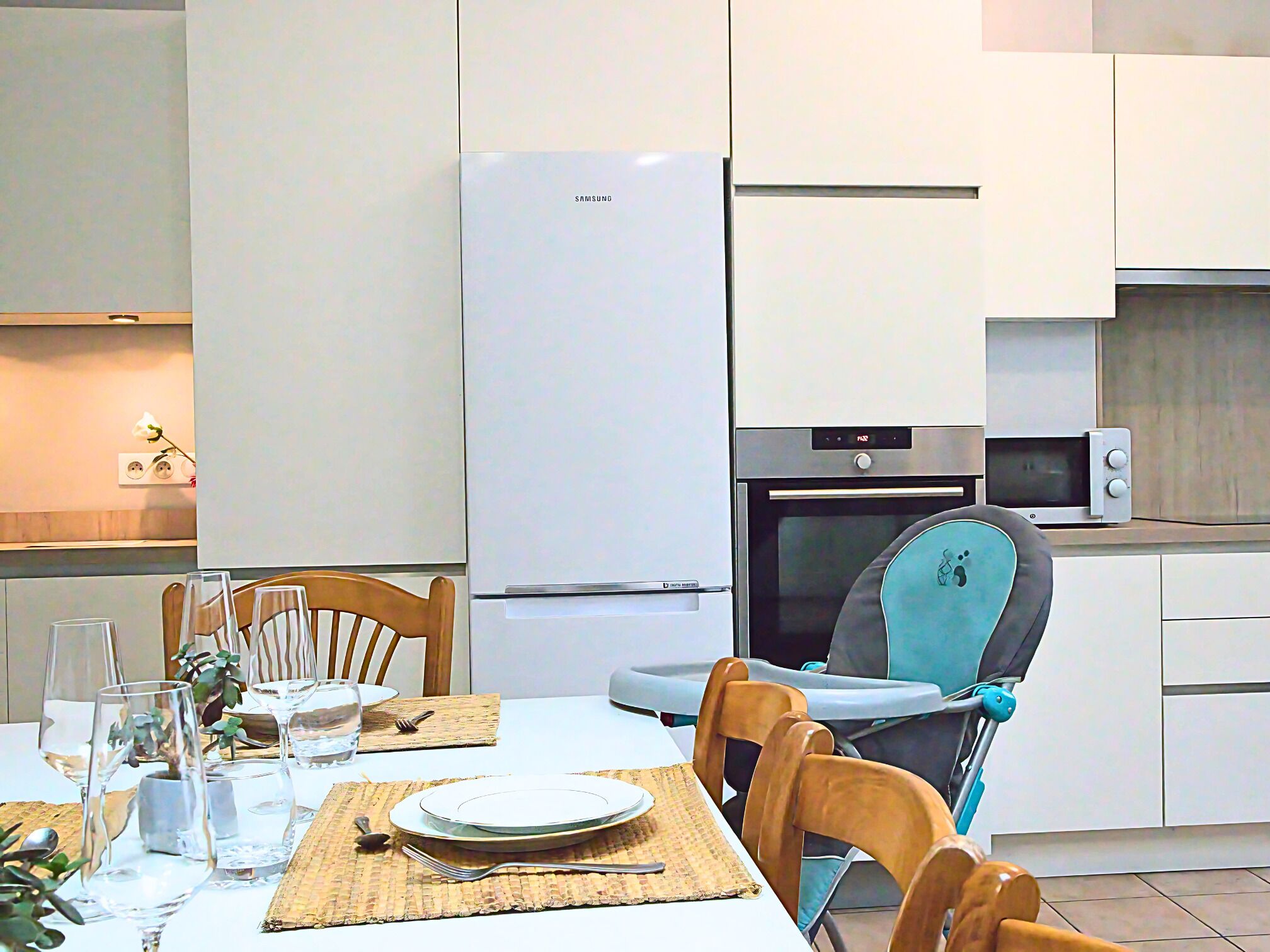 Een moderne keuken met een witte koelkast, magnetron en oven. Er staat een eettafel gedekt met borden, glazen en bestek. Een kinderstoel staat bij de tafel.