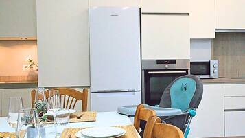 Een moderne keuken met een witte koelkast, magnetron en oven. Er staat een eettafel gedekt met borden, glazen en bestek. Een kinderstoel staat bij de tafel.