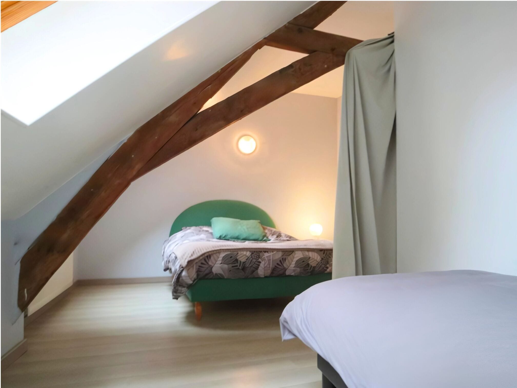 Una camera da letto con soffitto spiovente, una testiera verde, un letto con copripiumino fantasia, una lampada da comodino e una finestra con tende.