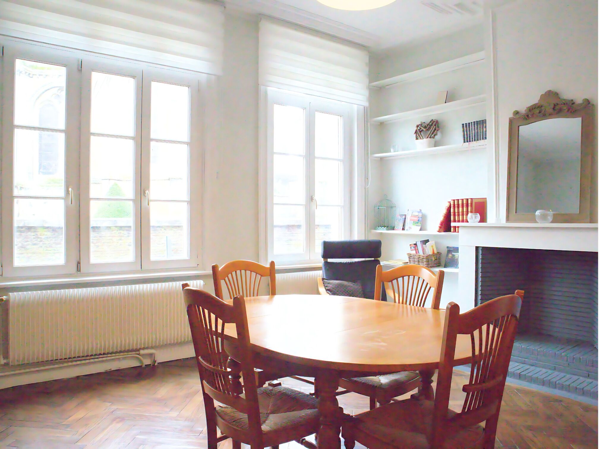 Un coin repas avec une table et des chaises en bois, de grandes fenêtres, une cheminée et une bibliothèque.