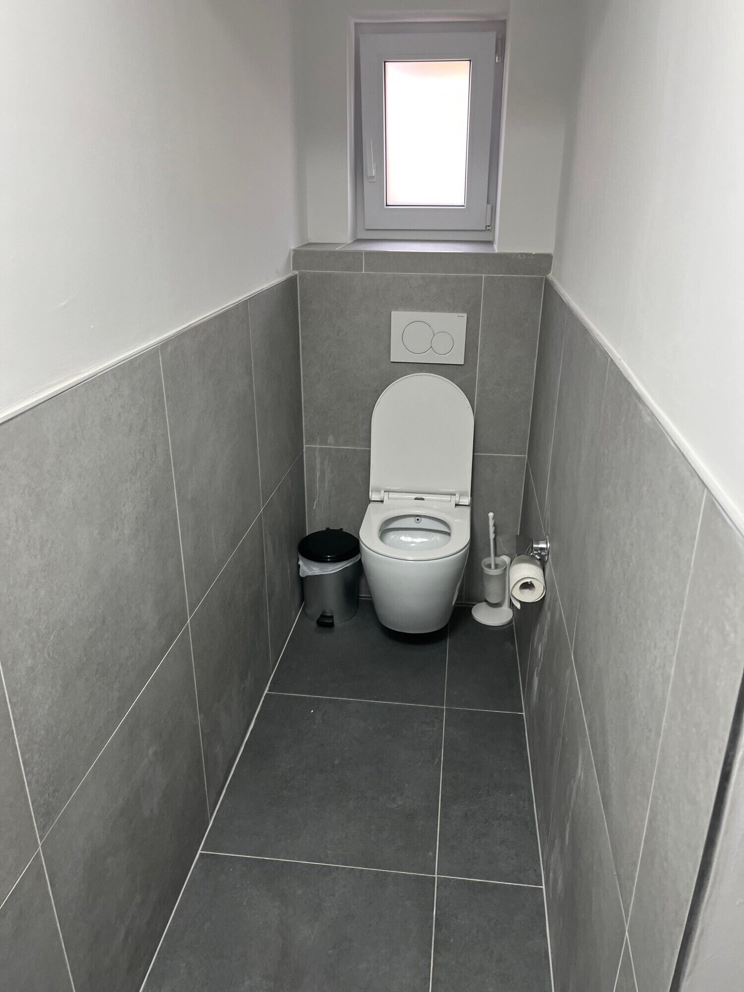 Een kleine, smalle badkamer met toilet, een prullenbak, een toiletrolhouder en een raam boven het toilet.