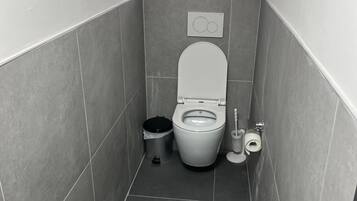 Een kleine, smalle badkamer met toilet, een prullenbak, een toiletrolhouder en een raam boven het toilet.