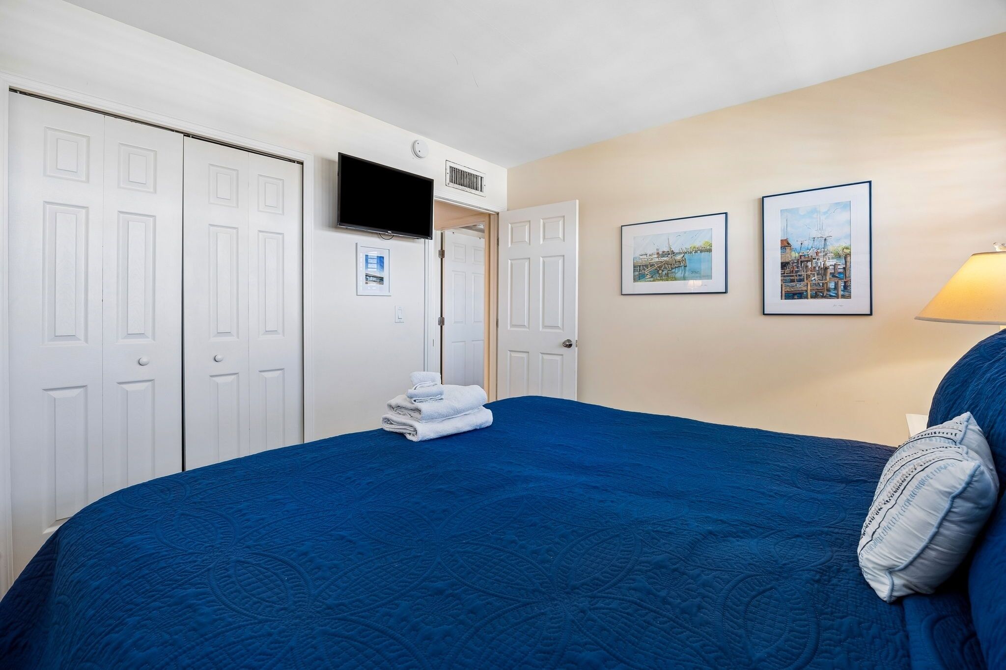 Ein Hotelzimmer mit einem großen Bett, einem weißen Kleiderschrank, einem Flachbildfernseher, zwei gerahmten Bildern an der Wand und einer Lampe auf der rechten Seite.