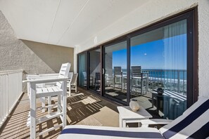 Ein Balkon mit weißen Stühlen und einem Tisch, eine Glastür, die auf eine Terrasse mit weiteren Sitzgelegenheiten führt, und Blick auf den Strand.