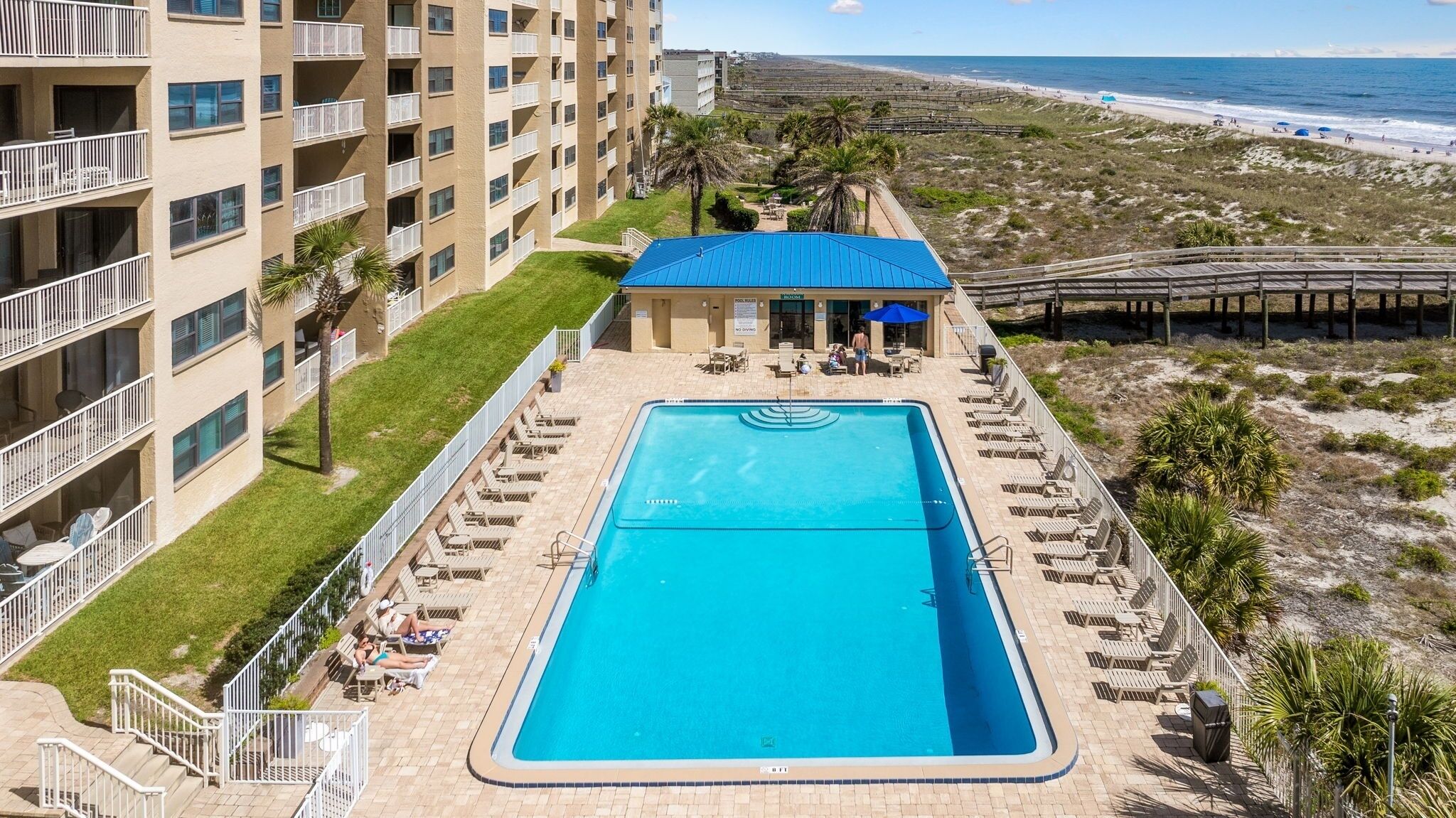 A hotel with a swimming pool, a beach view, and a covered area with umbrellas.