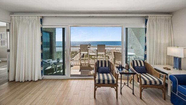 A room with a large sliding glass door leading to a balcony with beach views, two wicker chairs, a small table, and a blue sofa. - Cozy Ocean View Family Retreat - 3BR - Amelia by the Sea (Fernandina Beach)
