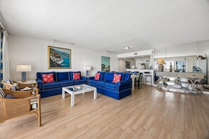 Uma sala de estar ampla com um sofá azul, uma mesa de centro branca, cadeiras de vime e uma cozinha ao fundo.