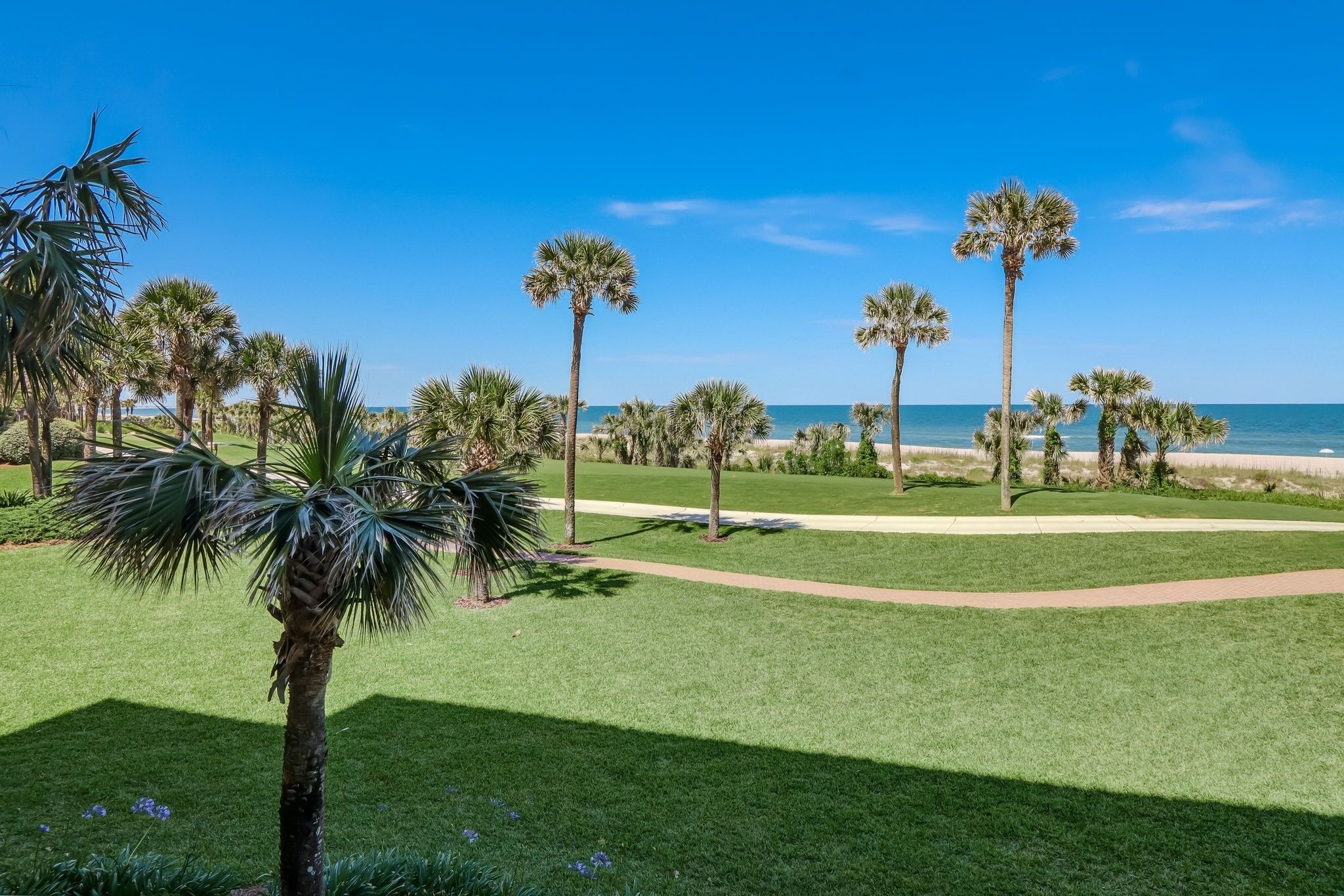 Un campo de golf bien cuidado con un sendero que lleva a la playa, rodeado de palmeras y un cielo azul despejado.