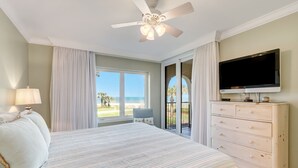 A bedroom with a bed, a ceiling fan, a TV, a dresser, and a view of the ocean through large windows. - Cozy Pet Friendly Condo by The Beach Omni Sea Dunes - 1616 (Fernandina Beach)