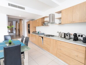 A modern kitchen with wooden cabinets, a gas stove, and various appliances. There is a dining area with a glass table and chairs. - Bormla Seafront 3 Bedroom Apartment (Birgu)