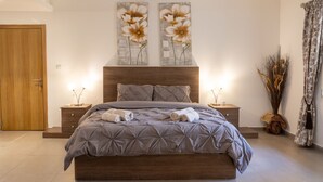 A modern bedroom with a large bed, wooden headboard, bedside tables, and two wall-mounted paintings. - Bormla Seafront 3 Bedroom Apartment (Birgu)