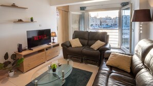 A modern living room with a leather sofa, a glass coffee table, a flat-screen TV, and a view of a cityscape through a large window. - Bormla Seafront 3 Bedroom Apartment (Birgu)