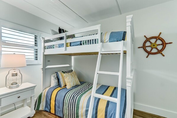 Une chambre avec un lit superposé, une couette à rayures, une table de chevet blanche avec une lampe et un gouvernail de bateau accroché au mur.