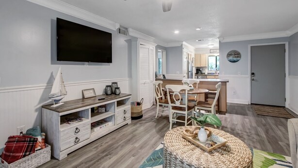 A modern living room with a flat-screen TV, wooden entertainment unit, and a dining area with a wooden table and chairs. - Fernandina Beach Hidden Gem Amelia Landings - 9F (Fernandina Beach)