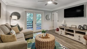 A living room with a sofa, ottoman, TV, and entertainment unit. - Fernandina Beach Hidden Gem Amelia Landings - 9F (Fernandina Beach)