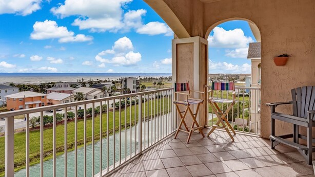 A balcony with a view of a river and houses, a ceiling fan, and a small table with a chair. - Top Floor Beach View Condo Ocean Park 409 (Fernandina Beach)