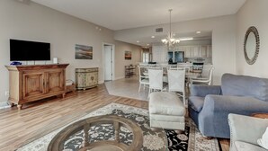 A spacious living room with a large flat-screen TV, a wooden cabinet, and a dining area with white chairs and a chandelier. - Top Floor Beach View Condo Ocean Park 409 (Fernandina Beach)
