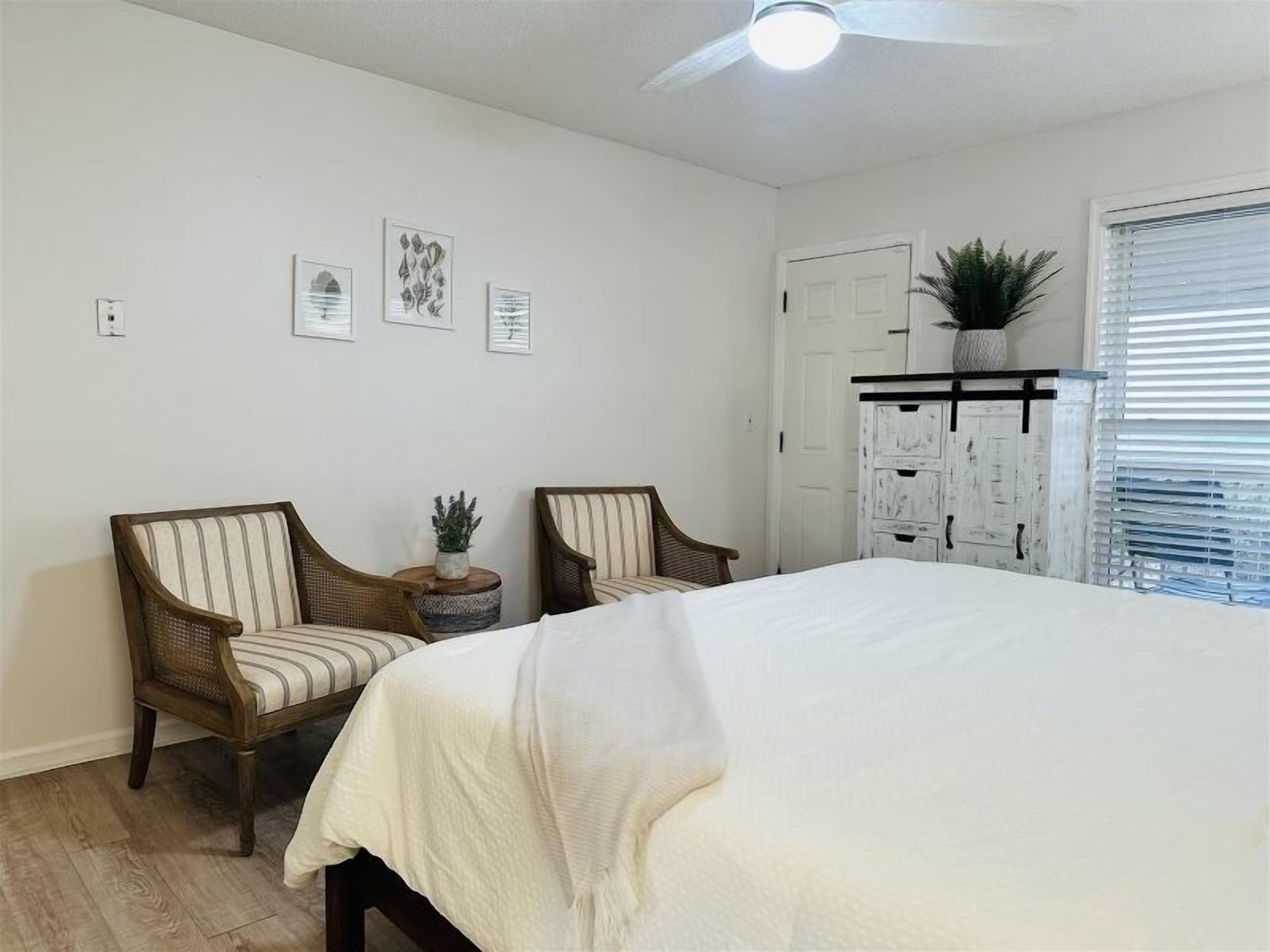 A bedroom with a large bed, two upholstered chairs, a ceiling fan, a white wardrobe, and framed artwork on the wall.