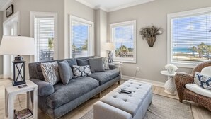 A living room with a grey sofa, a white side table, a lamp, a wicker chair, and large windows with blinds. - Luxury Private Beachfront Condo w/ Parking Garage & Stunning Views (Fernandina Beach)