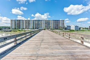 A wooden boardwalk leading to a large hotel with multiple floors and balconies, surrounded by greenery and a clear sky. - Beachfront Family Condo w/ Pool, Fishing, and Pier Access Amelia by the Sea (Fernandina Beach)