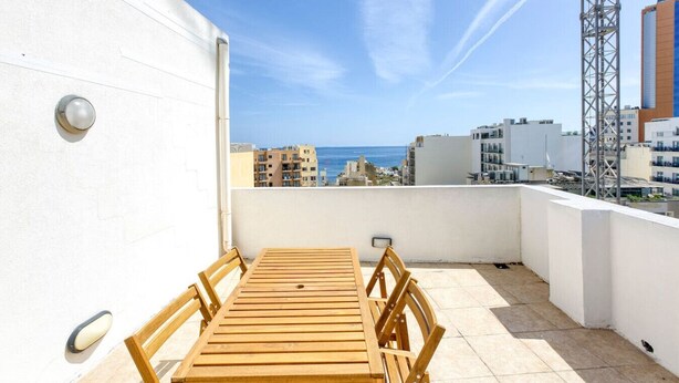 Une terrasse sur le toit, avec une table et des chaises en bois, un mur blanc et une vue sur la ville et la mer.