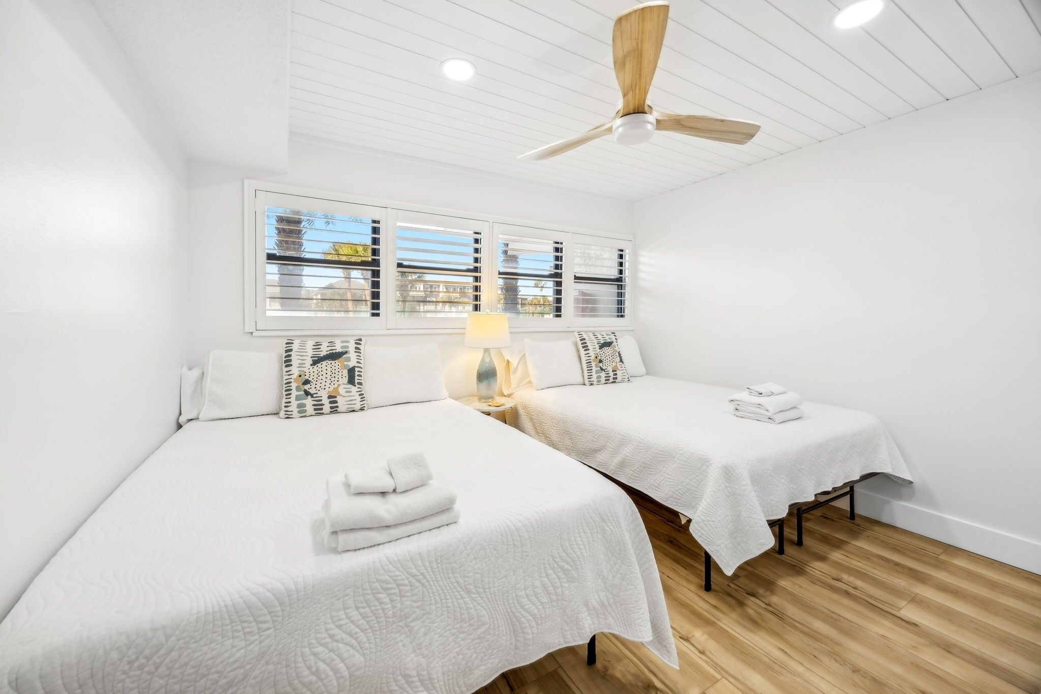 Une chambre avec deux lits, un ventilateur de plafond, une fenêtre avec des stores et une table de chevet avec une lampe.
