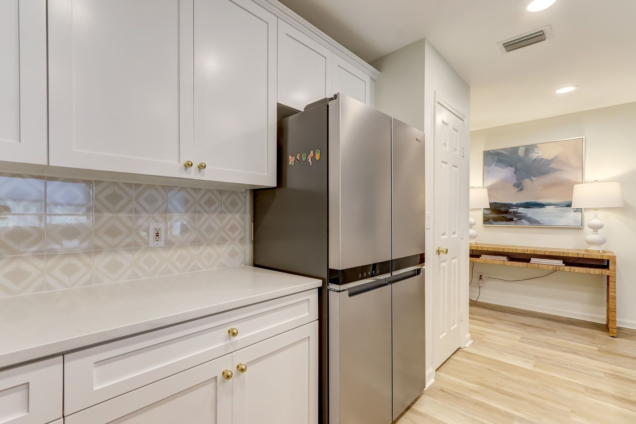 A modern kitchen with white cabinets, a stainless steel refrigerator, and a wooden console table with a lamp.