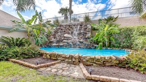 Un patio trasero con piscina, cascada de piedra y abundante vegetación.